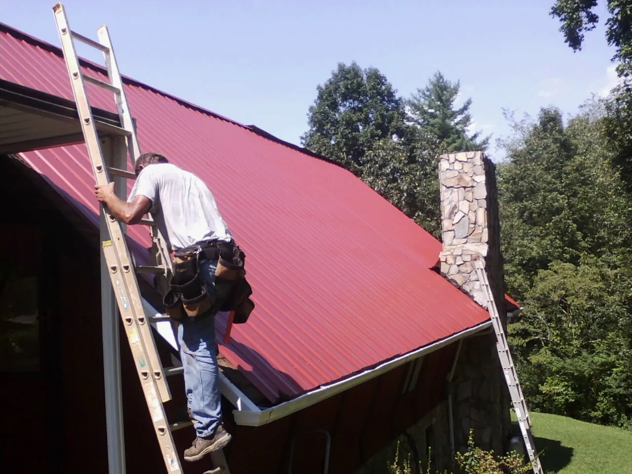 Roofer on a ladder beside a red metal roof and stone chimney — Soffit & Fascia Repair by Roofing Brothers in Virden